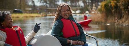 Two people wearing red life jackets smile and relax on a paddle boat, gliding across a calm lake, with reeds, trees, and another small red boat in the background.