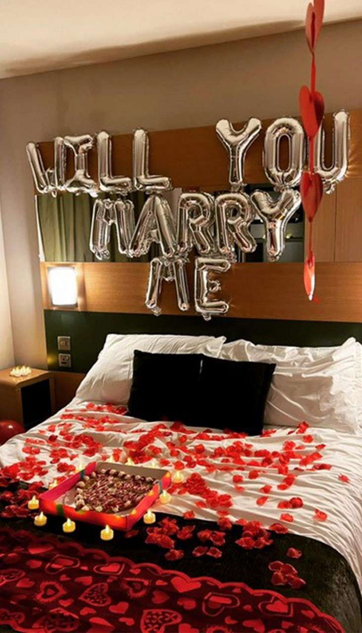 Bed decorated with red rose petals, small candles, and a gift tray, in a hotel room where silver balloons spell "WILL YOU MARRY ME" above the headboard.