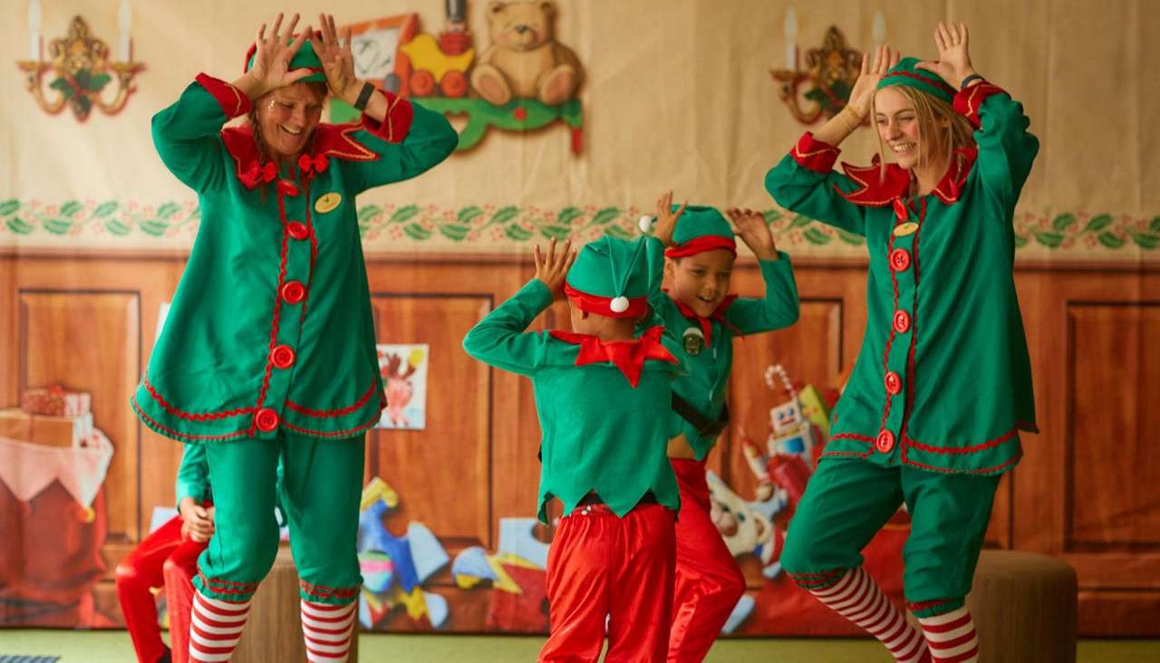 People in green elf costumes dance with hands posed like antlers, joined by two children, in a festive indoor room with decorations, a wood-paneled backdrop, and candy-cane striped socks.