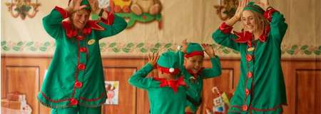 Four people in green elf costumes raise hands like antlers, smiling and playing together in a festively decorated room with wooden paneling, garlands, and holiday crafts on the walls.