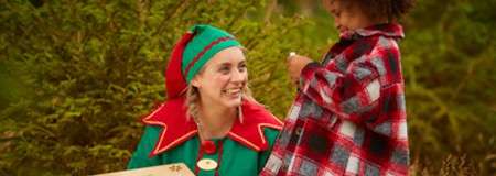 Elf-costumed adult kneels and smiles at a child, presenting a decorated box. The child, in a red plaid poncho, examines something in hand amid evergreen trees outdoors.