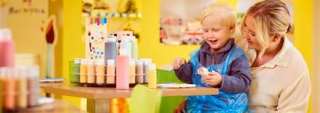 Child paints a small object while an adult assists, smiling. Colorful paint bottles line the table in front. Context: bright, playful art studio with yellow walls and children’s artwork.