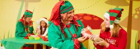 Elf-costumed adult hands a wrapped gift to a child in a striped elf hat; behind them, two people craft at a table; brightly colored, cartoonish holiday workshop backdrop.
