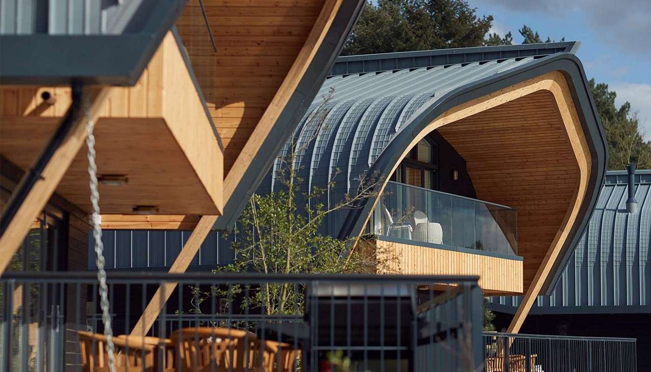 Curved, timber-lined balconies project from modern buildings, sheltering glass-railed terraces with chairs; ribbed metal roofs sweep overhead. In back, more units, trees, and blue sky complete the residential setting.