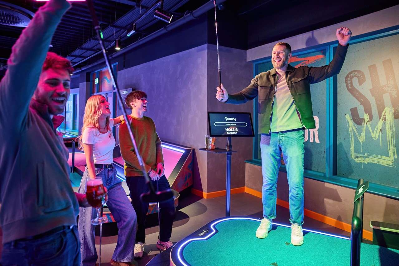 Man raises golf club in celebration while standing on a glowing mini-golf green; friends laugh nearby in a neon-lit indoor venue with a screen reading: Puttery HOLE 6 FAIRWAY.