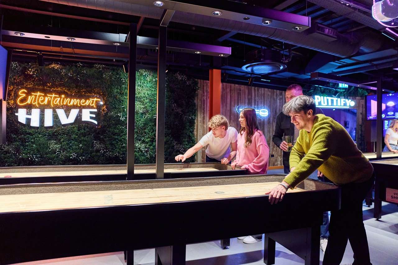 Friends shuffle pucks on a long shuffleboard table, smiling and leaning forward, within a neon‑lit arcade setting. Visible signs read: “Entertainment HIVE” and “PUTTIFY”.