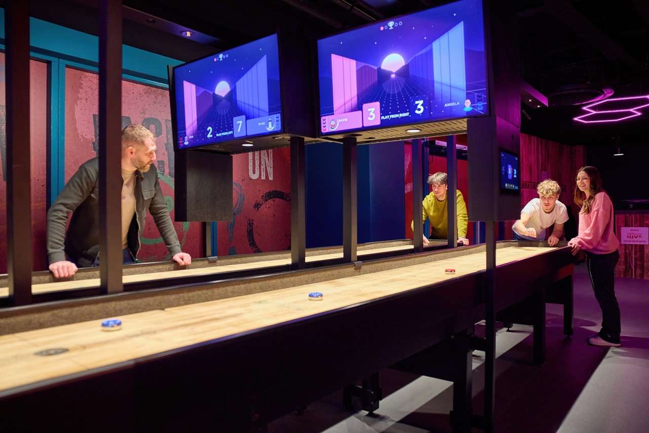 Players slide shuffleboard pucks across a long wooden table, competing under overhead displays in a neon-lit arcade bar. Text: “2,” “7,” “3,” “3,” “PLAY FROM RIGHT,” “DAVID,” “ANGELA,” “ON,” “GIVE.”
