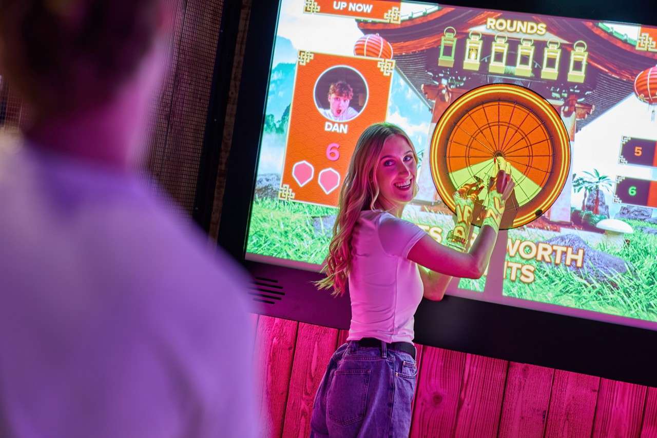 Shuffleboard players push red pucks along a long wooden table in a neon-lit game bar; overhead scoreboards show: DAVID 6, PLAY FROM LEFT; ANGELA 0; DAN 1, PLAY FROM RIGHT; SHUFL 4.