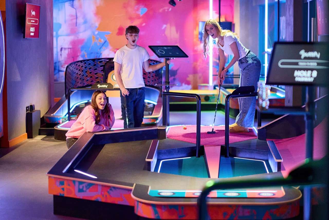 Golfer putts on a bright mini-golf hole, while two friends watch and react, in a neon-lit, colorful indoor arcade. Text: "HOLE 5"; "HOLE 8".