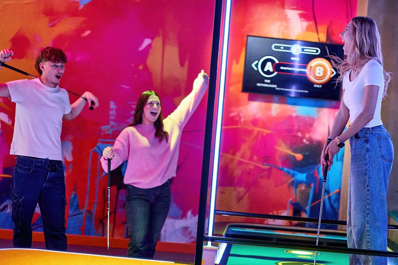 Three people with mini-golf putters celebrate; one prepares to putt, two raise arms. Neon-lit indoor mini-golf course surrounds them, with glowing rails, mural, and a screen displaying “A” and “B”.