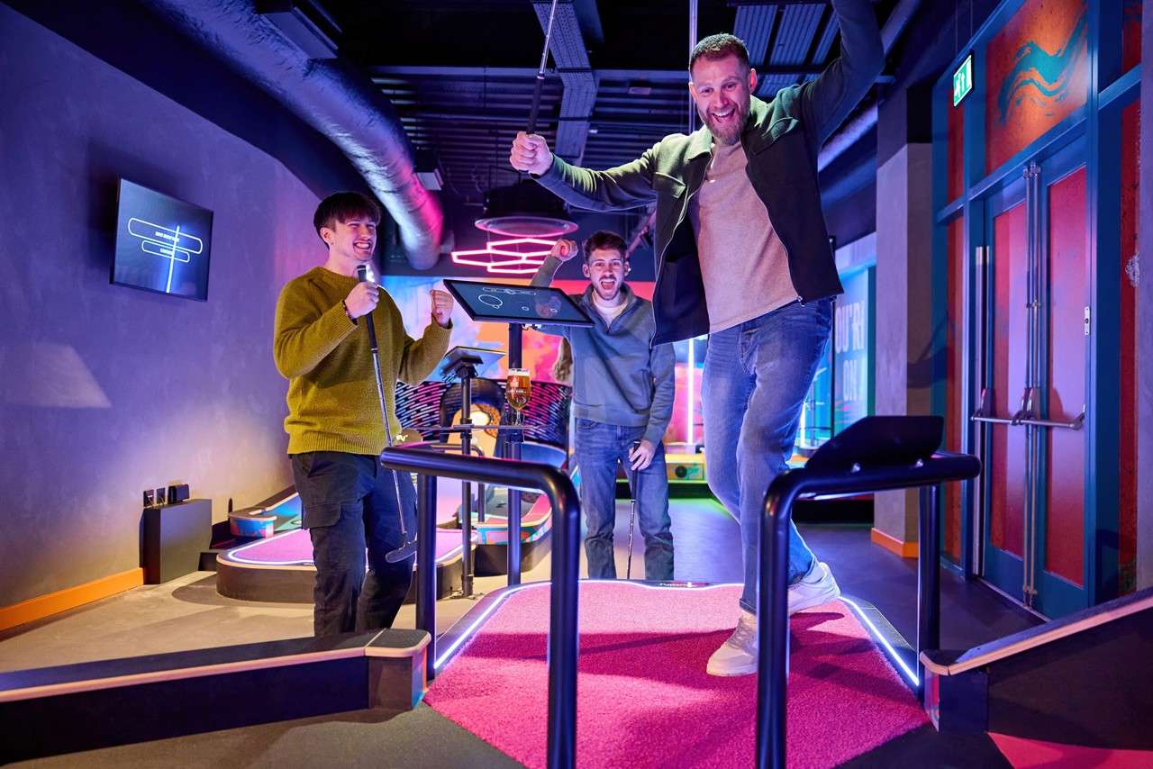 Man celebrates a shot, raising arms while stepping off a pink mini-golf platform; two companions cheer with putters. Neon-lit indoor course features rails, screens, spiral lights, and a beer glass. Text: EXIT.