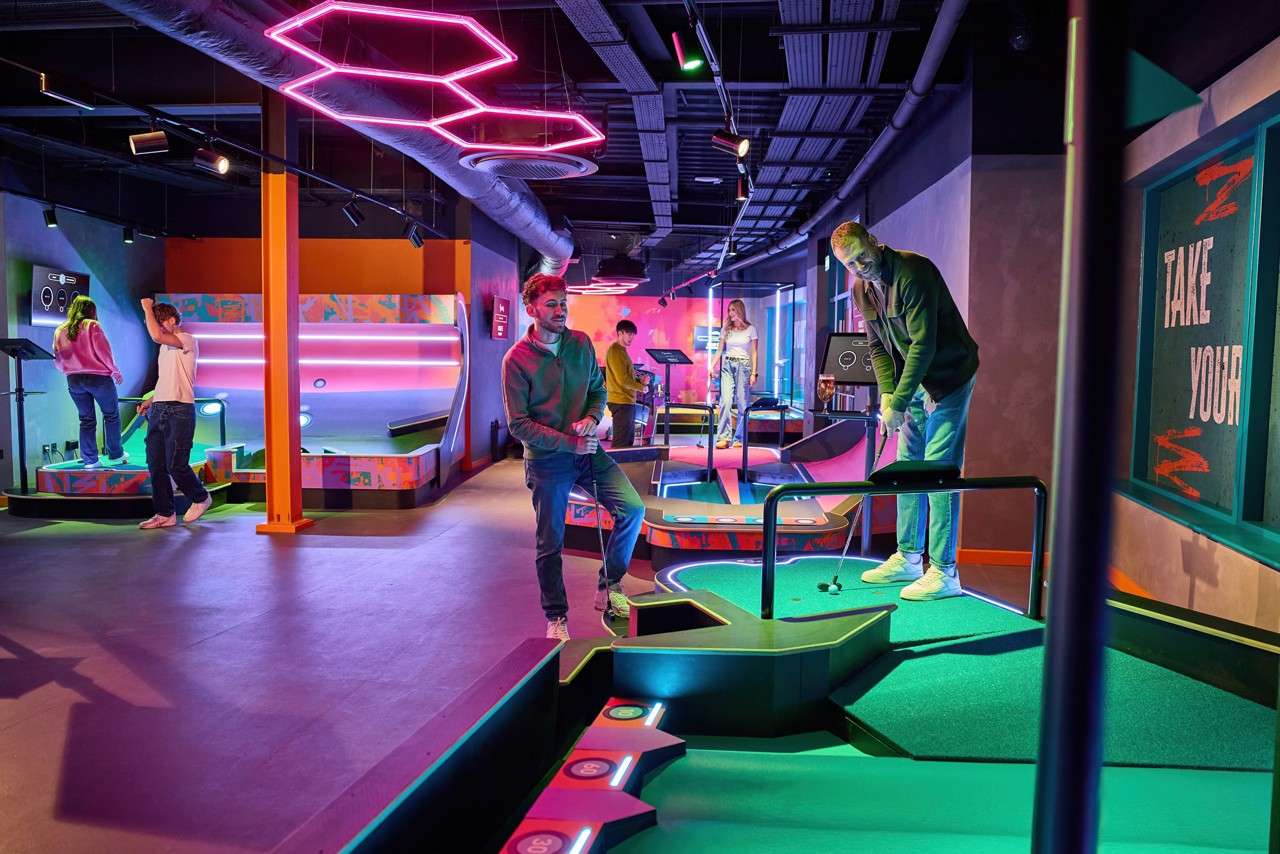 People putt golf balls, navigating ramps and holes, inside a neon-lit indoor mini-golf venue with colorful obstacles and hexagonal lights; text: TAKE YOUR; 30, 40, 50.