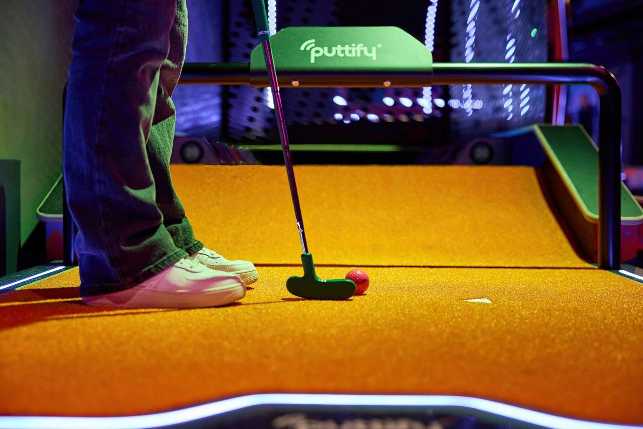 Putter addresses a red mini-golf ball; player stands on orange turf. Indoors, a ramp leads to a goal under a sign reading “puttify” with neon lighting.