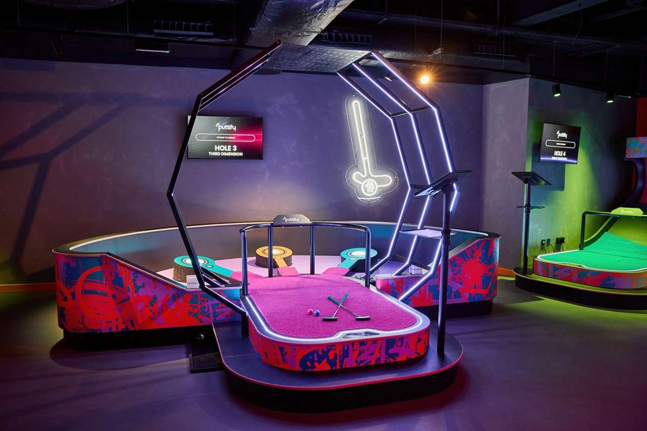 Mini-golf platform holds crossed putters and balls, awaiting play; an angular neon arch frames it within colorful lanes and glowing screens in a dark indoor arcade. Text: HOLE 3 THIRD DIMENSION; HOLE 4.