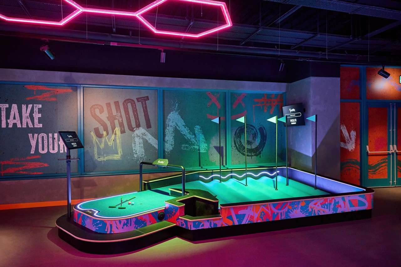 Mini-golf hole awaits play; balls and a putter rest on turf. Neon lighting and graffiti-style walls surround the course with flags and a screen. Text: TAKE YOUR SHOT; HOLE 8.