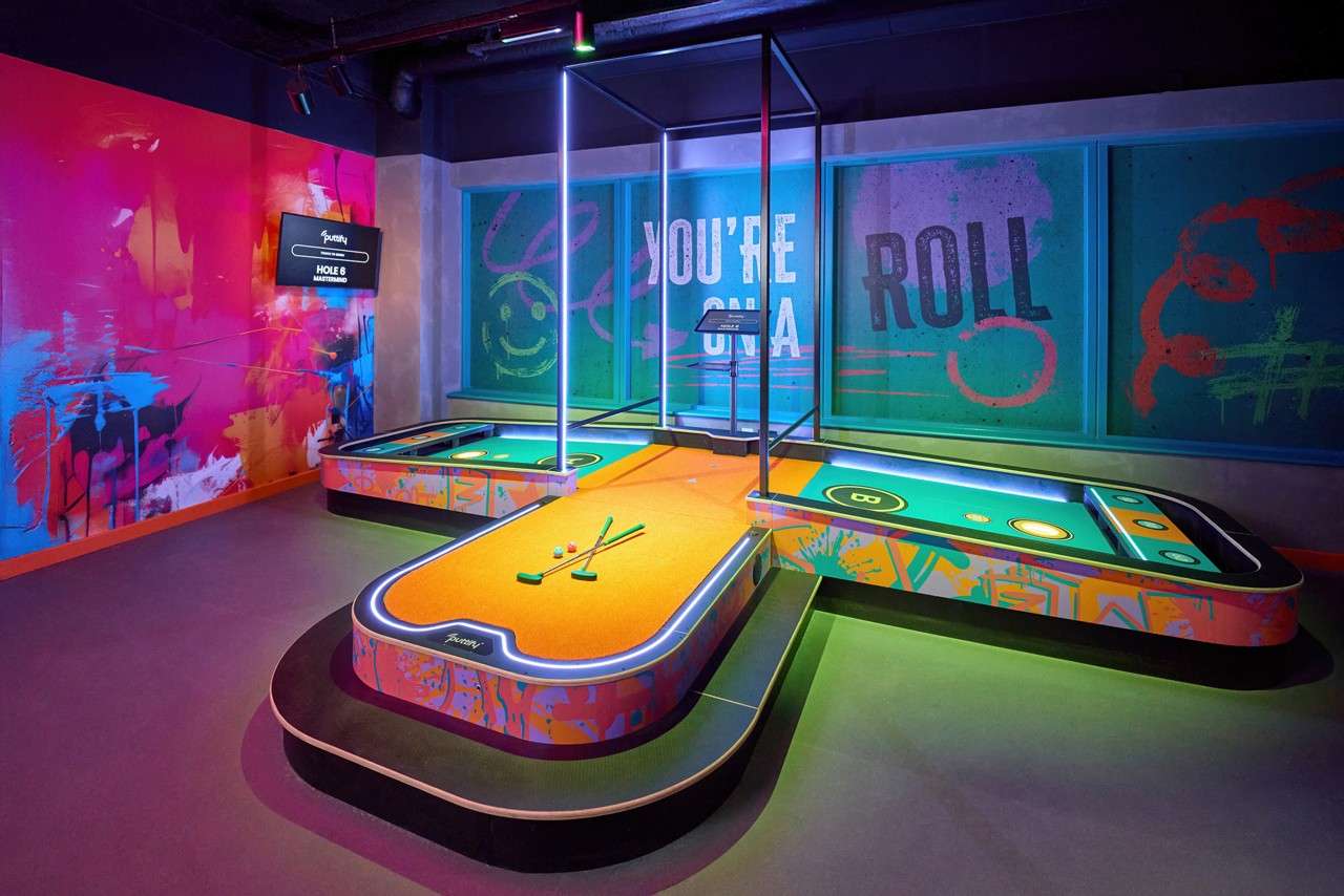 Mini‑golf course sits glowing, clubs and balls arranged on an orange start platform, in a neon, graffiti‑styled indoor venue. Text: YOU’RE ON A ROLL; HOLE 8; 8.