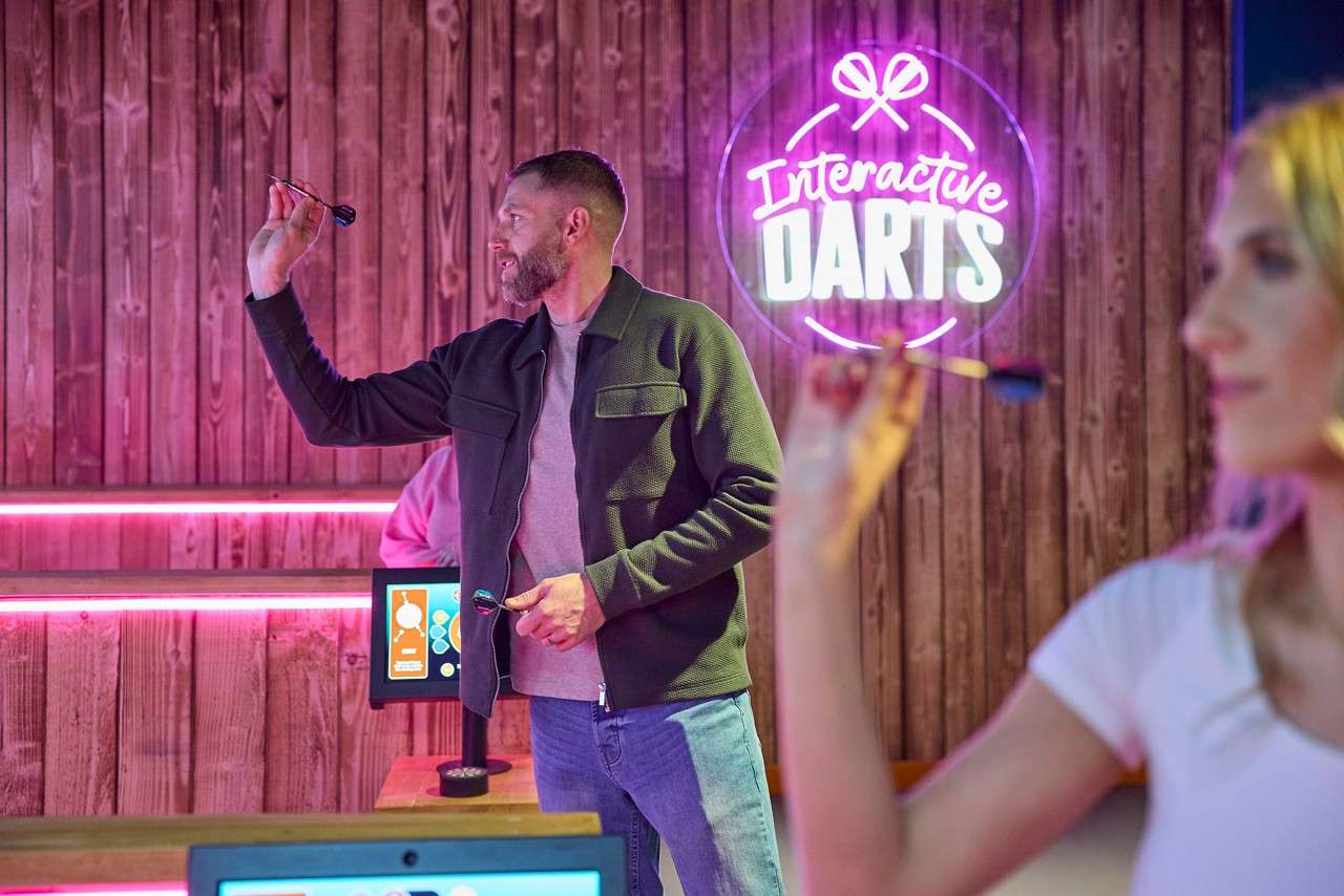 Man aims a dart, preparing to throw; woman in foreground also holds a dart. Neon sign reads: Interactive DARTS. Context: wood-paneled, neon-lit gaming venue with touchscreens.
