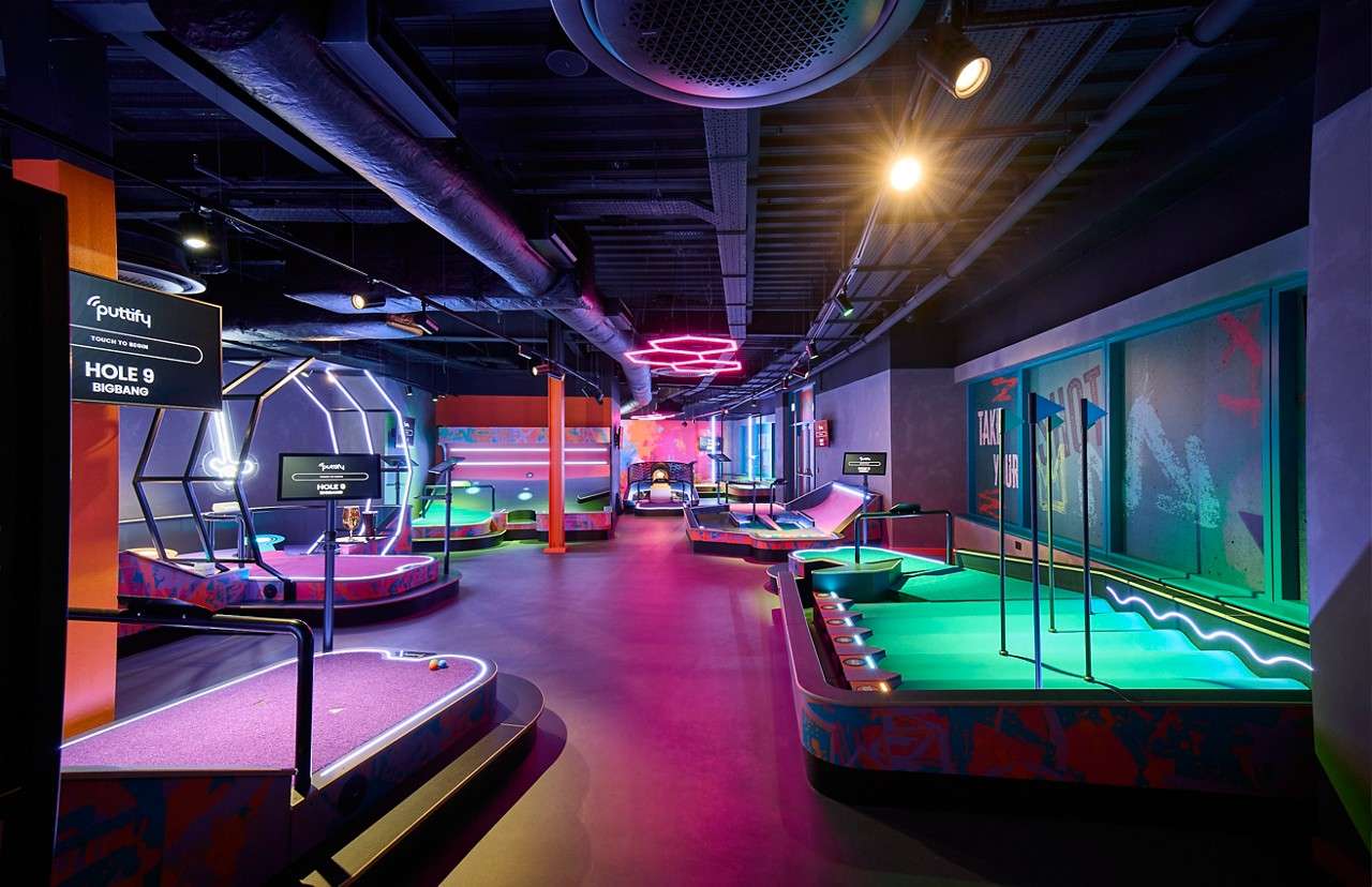 Mini-golf courses glow, inviting play, in a neon-lit indoor arcade with geometric obstacles and digital score screens. Visible text: puttify; TOUCH TO BEGIN; HOLE 9; BIGBANG; TAKE YOUR SHOT.