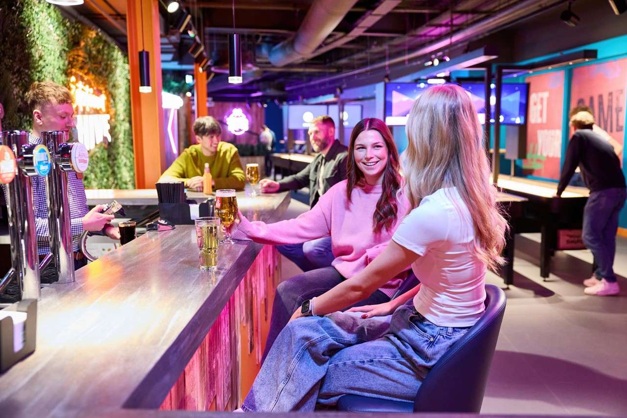 Two women sit at a bar, chatting and drinking beer, while others socialize and play shuffleboard; neon, colorful lighting fills a modern arcade venue. Text: GET, GAME.
