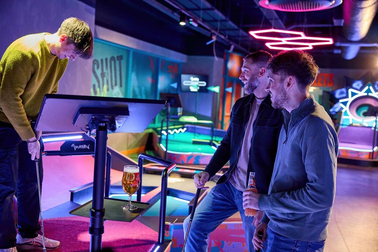 Golfer bends to putt while two friends watch and hold drinks in a neon-lit indoor mini-golf venue, beside a touchscreen stand holding a beer. Text visible: SHOT; puttify.