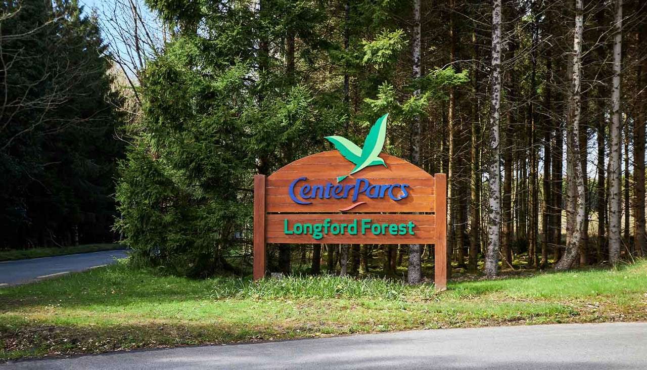 Wooden roadside sign displays “Center Parcs” and “Longford Forest,” standing at a forest entrance beside a paved road, surrounded by tall evergreen trees.