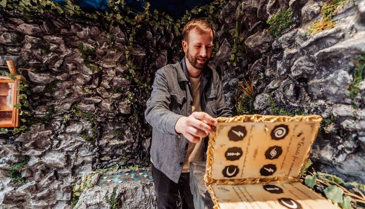 Man opens an ornate, oversized book, examining pages with round icons—lightning bolt, tree, crescent moon, spirals—while standing in a faux-stone, vine-covered room with a small water feature. Text on pages is unreadable.