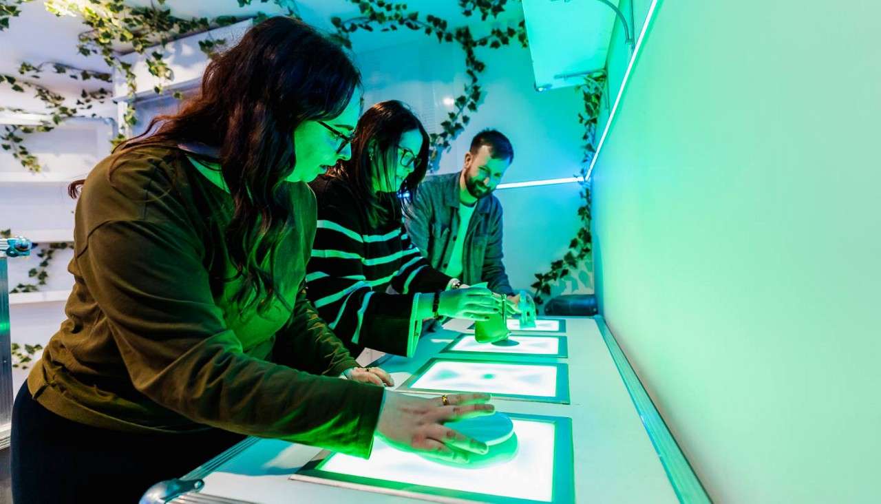 Three participants press glowing rectangular touchpads and move puck-like devices on a counter, concentrating, in a green-lit interactive exhibit room with vines on walls and shelves.