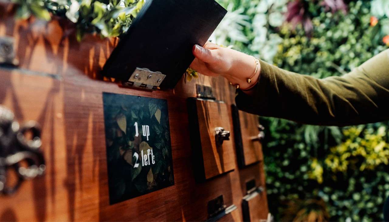 Hand opens a hinged wooden panel on a puzzle wall; neighboring square displays text: 1 up 2 left; surrounding area features multiple latches and leafy greenery in the background.