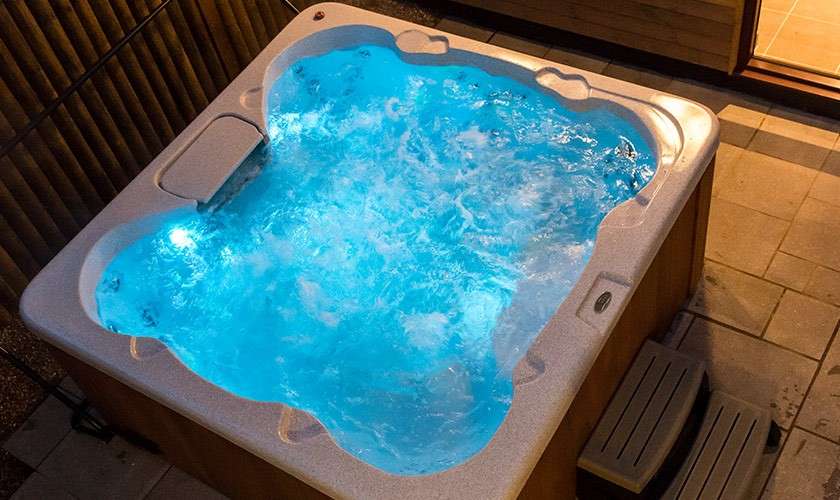 Hot tub bubbles vigorously, blue-lit water swirling. It sits on an outdoor patio beside tiled flooring, a wooden privacy screen, and small entry steps, viewed at night from above.