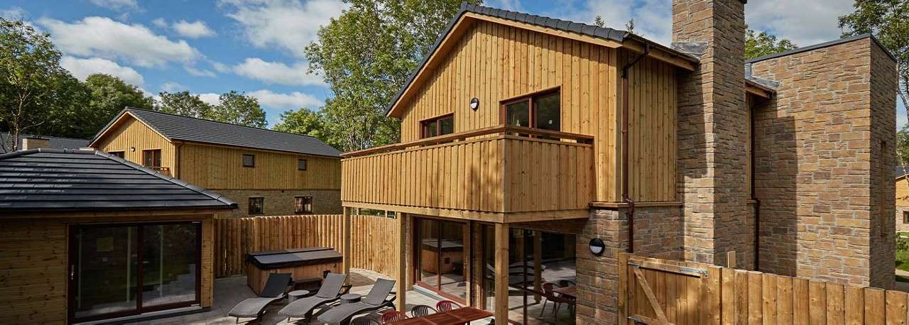 Timber-clad cabin overlooks a fenced patio, featuring lounge chairs, dining table, and hot tub. Surrounding buildings echo the wood-and-stone style, while leafy trees and blue sky frame the quiet setting.