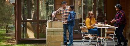 Family barbecues on a patio; one tends the grill while a child assists; two others chat at a table; trees and a glass-walled cabin form the background.