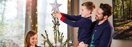 Child places a silver star atop a Christmas tree while an adult lifts him; another child watches. Indoors near large windows, warm string lights glow and ornaments decorate the tree.