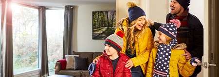 Four bundled-up people enter a bright living room, smiling and talking; sunlight streams through large windows, with a couch and a framed forest photo in the background.