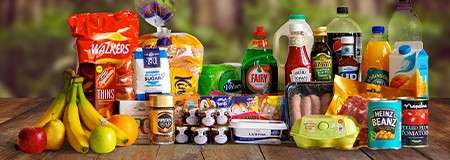Assorted groceries sit arranged on a wooden table outdoors, including crisps, bread, milk, cereal, washing-up liquid, ketchup, sausages, eggs, fruit, and drinks. Visible text: WALKERS, THINS, FAIRY, HEINZ, BEANZ.
