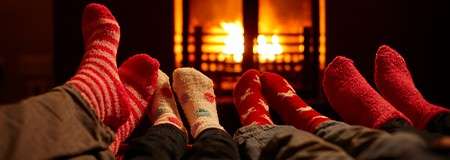 Feet in colorful, patterned winter socks rest close together, warming by a glowing fireplace. Soft orange firelight illuminates a dim room, with multiple people reclining indoors during cold weather.