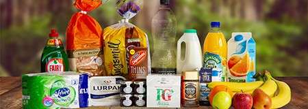 Assorted groceries sit arranged on a wooden table; products include fruit, bread, milk, juice, tea, butter, detergent, and toiletries. Visible text: FAIRY, Warburtons, Kingsmill, THINS, LURPAK, PG, Orange.
