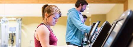 Two adults walk on treadmills, focusing on the control panels, in a well-lit gym with beige walls and equipment in the background.