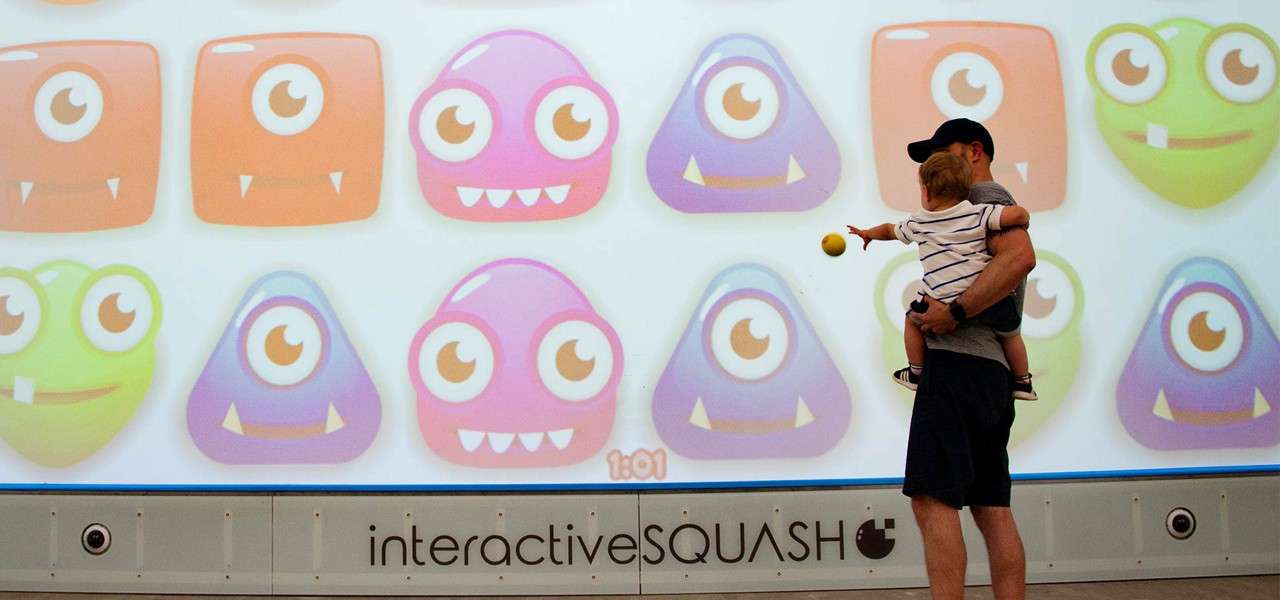 Child, held by an adult, tosses a yellow ball toward a large screen of colorful one-eyed cartoon monsters in an interactive court. Text: interactiveSQUASH, 1:01
