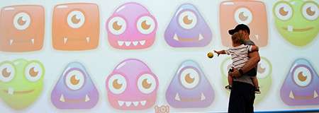 Adult holding a child points toward a large screen displaying rows of colorful, one-eyed cartoon monster icons with fangs. They stand in front of the playful projection indoors.