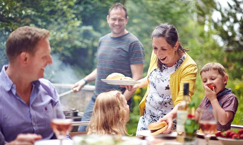 Group at outdoor table—woman serves buns, boy eats strawberry, girl looks up, man grills behind, another adult chats—set in green backyard garden with drinks, plates, and barbecue smoke.