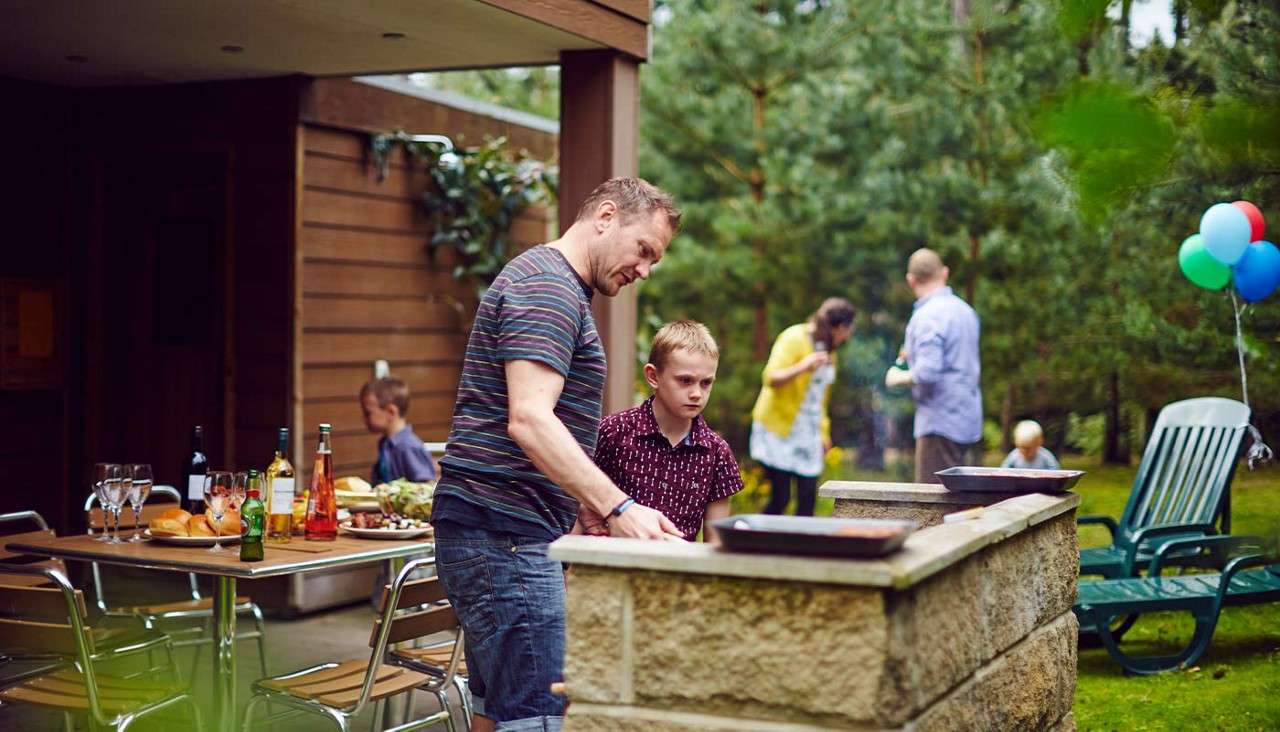 Man and boy grill at a stone barbecue as others chat nearby. In a wooded backyard, a table holds drinks and dishes, lawn chairs, and balloons indicating a casual celebration.