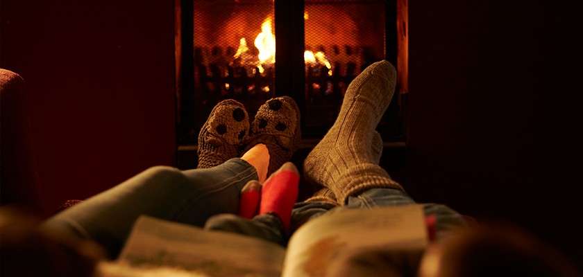 Two people’s socked feet rest together, facing a glowing fireplace; one holds an open book. They lounge on a couch, warming by the fire in a dim, cozy room.