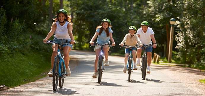 Four cyclists ride forward, smiling, wearing helmets; two adults and two children pedal along a sunlit paved path through a green wooded park, dappled light and shadows on the road.