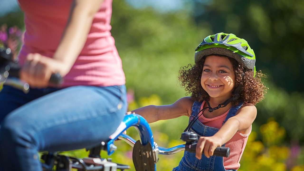 Smiling child wearing a green helmet rides a bike, following an adult cyclist, in a sunny park with greenery and blurred flowers in the background.