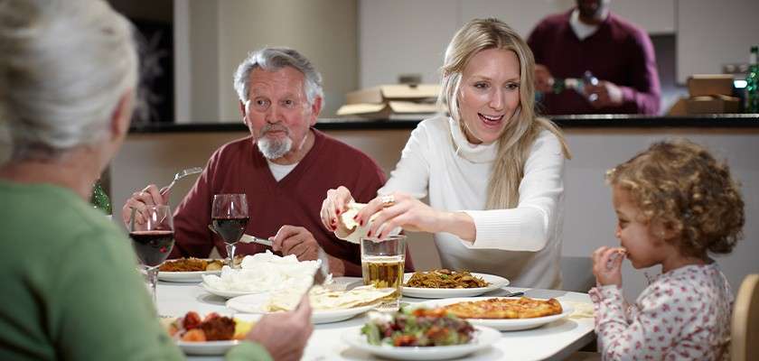 Family at dinner eats and chats, passing bread and sharing pizzas, salad, and dishes; glasses of wine and beer on table; child snacks; kitchen background with another person preparing food.