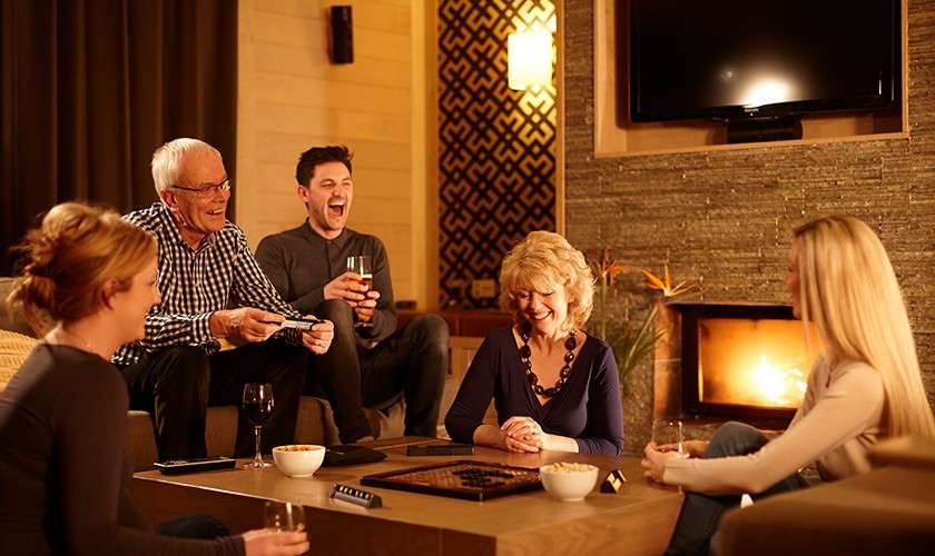 Group of five adults laugh while playing a board game, seated around a coffee table in a warmly lit living room with a fireplace, wine glasses, and snacks.