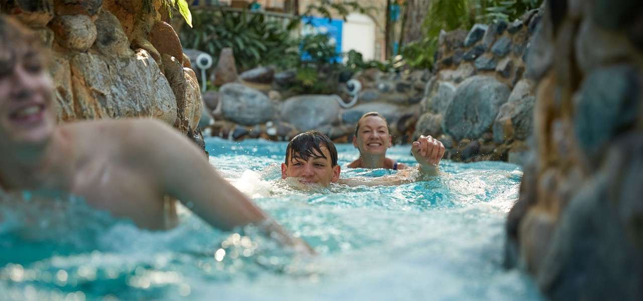 Three people swim and float forward, smiling, as churning blue water pushes them along a narrow, rock-lined lazy river surrounded by tropical plants inside an indoor water park.