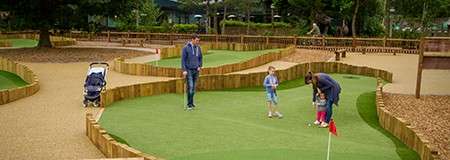 Family playing mini-golf: an adult helps a child putt while another adult and child watch; stroller nearby. Context: landscaped course with wooden borders, winding greens, trees in background.