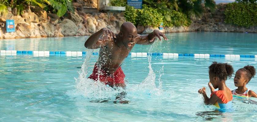 Man splashes playfully, raising water with both hands, while two children watch and laugh in a shallow outdoor pool surrounded by rocks, plants, and lane-marking floats.
