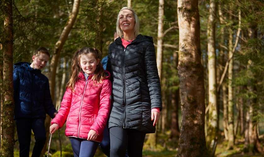 Family walkers stride together; an adult in a black coat and two children in bright jackets walk and smile, holding a stick, among tall trees on a sunlit forest trail.